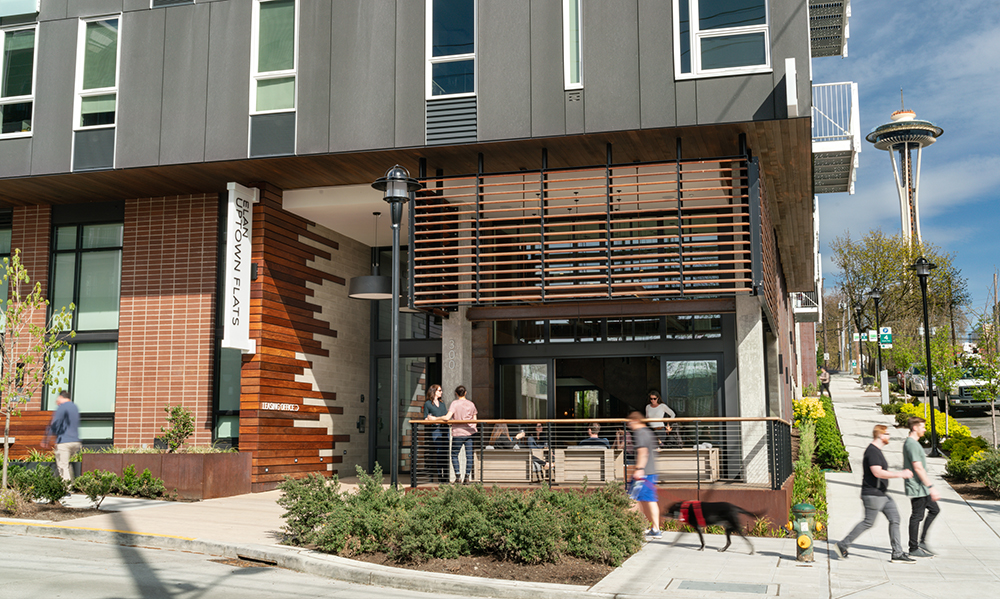 photo of the porch at uptown flats with people sitting around the fire pit and people walking on the sidewalk and the space needle in the background