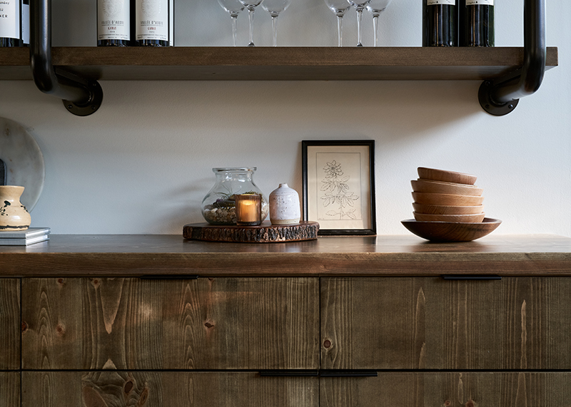 photo of a decorated wood wine cabinet with wine bottles, a low lit candle, and wooden bowls