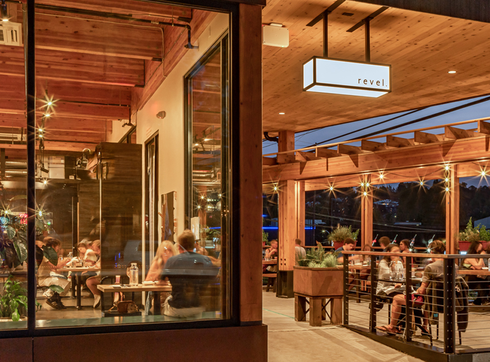 photo of the exterior of revel restaurant in seattle at dusk with people sitting outside on the patio and people inside dining through the window