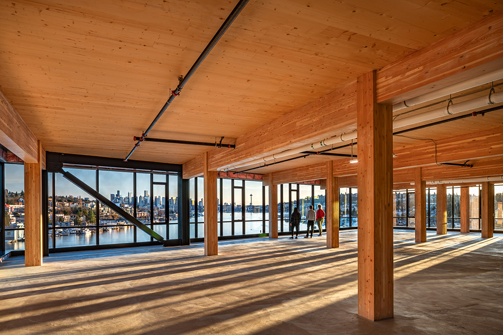 photo of the inside of northlake commons highlighting the mass timber ceilings and columns. three people look outside the windows overlooking lake union with the seattle skyline in the backdrop