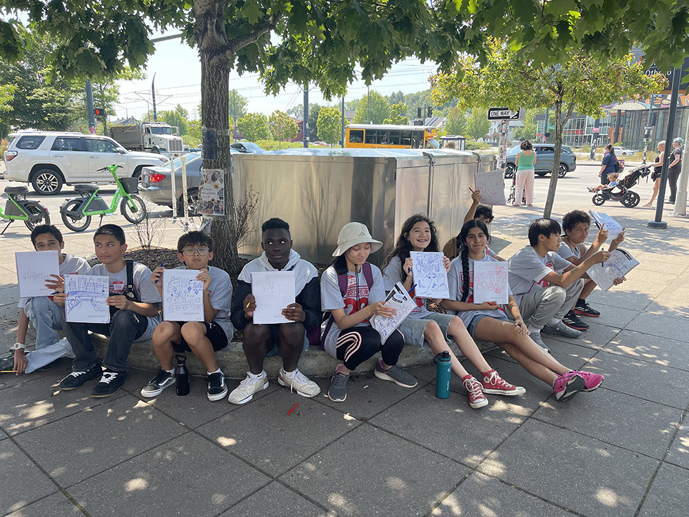 photo of students holding up their sketches outside, sitting on a curb under a tree